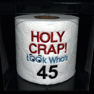 May include: A white toilet paper roll with red and blue embroidery that says "HOLY CRAP! LOOK WHO'S 45". The roll is in a clear plastic box.