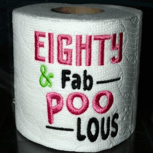 May include: A white toilet paper roll with the text "EIGHTY & Fab-POO-Lous" embroidered in pink, green, and black thread.