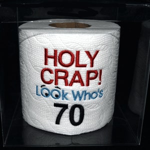 May include: A white toilet paper roll with the text "HOLY CRAP! LOOK Who's 70" embroidered on it in red and blue thread. The toilet paper roll is in a clear plastic box.