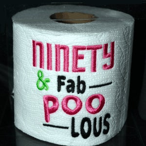 May include: A white toilet paper roll with the text "Ninety & Fab-Poo-Lous" embroidered in pink, green, and black thread.
