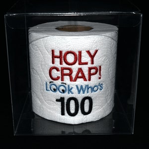 May include: A white toilet paper roll with embroidered text in red and blue that reads "HOLY CRAP! Look Who's 100" inside a clear plastic box. The text is centered on the roll, with the number 100 in black.