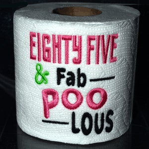 May include: A roll of white toilet paper with the embroidered text "EIGHTY FIVE & Fab-POO-LOUS" in pink, green, and black thread. The text is centered on the roll.