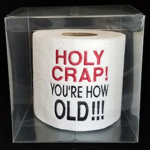 May include: A white toilet paper roll with the text "HOLY CRAP! YOU'RE HOW OLD!!!" in red and black embroidery. The roll is inside a clear plastic box.