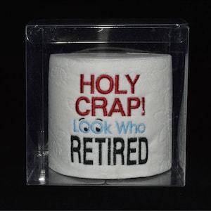 May include: A white toilet paper roll with the text "HOLY CRAP! LOOK WHO RETIRED" embroidered on it. The text "HOLY CRAP!" is in red, "LOOK WHO" is in blue, and "RETIRED" is in black. The toilet paper roll is in a clear plastic box.