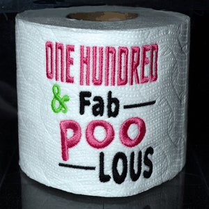 May include: A white toilet paper roll with the text "One Hundred & Fab-Poo-Lous" embroidered on it in pink, green, and black thread.