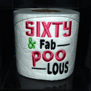 May include: A white toilet paper roll with the text "SIXTY & Fab-POO-LOUS" embroidered on it in pink, black, and green thread. The roll is in a clear plastic container.