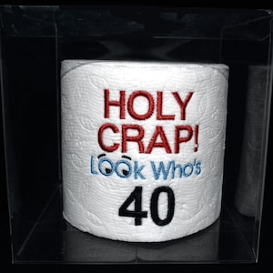May include: A white toilet paper roll with the text "HOLY CRAP! LOOK Who's 40" embroidered on it. The toilet paper roll is in a clear plastic box.