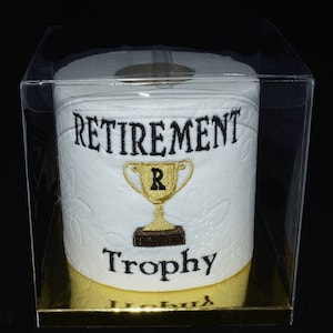 May include: A white toilet paper roll with black embroidery that reads "Retirement Trophy" and a gold trophy with the letter "R" on it. The toilet paper roll is in a clear plastic box with a gold base.