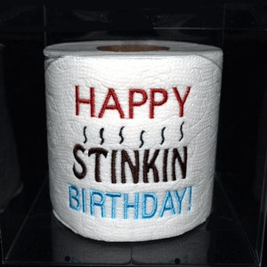 May include: A white toilet paper roll with the embroidered text "HAPPY STINKIN BIRTHDAY!" in red, brown, and blue. The roll is displayed inside a clear, square case against a black background.