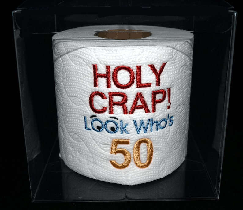 Puede incluir: Un rollo de papel higi&eacute;nico blanco con el texto bordado "HOLY CRAP! LOOK WHO'S 50" en hilo rojo, azul y dorado. El papel higi&eacute;nico est&aacute; encerrado en una caja de pl&aacute;stico transparente, sobre un fondo negro.
