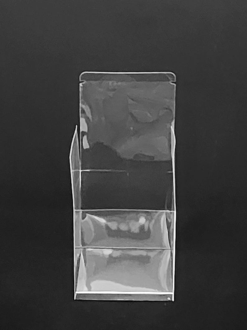 Puede incluir: Un expositor de pl&aacute;stico transparente con tres bolsillos escalonados. El soporte es rectangular con una parte superior plegada y est&aacute; colocado sobre un fondo negro. Los bolsillos est&aacute;n dise&ntilde;ados para guardar y exhibir art&iacute;culos.