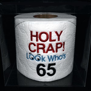 May include: White toilet paper roll with the embroidered text "HOLY CRAP! LOOK WHO'S 65" in red, blue, and black. The roll is encased in a clear plastic box, set against a black background.