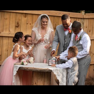 May include: A wedding ceremony with a bride and groom, two flower girls in pink dresses, and two ring bearers. The bride and groom are pouring sand into a glass jar with the words "Love & Happily Ever After" on it. The ceremony is taking place outdoors in front of a wooden fence.