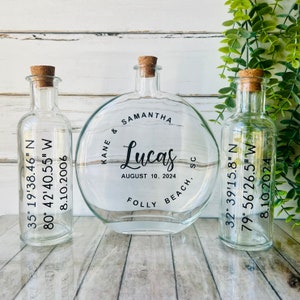 May include: Three clear glass bottles with cork stoppers. The central bottle is round and inscribed with "Samantha & Lucas" and the date "August 10, 2024". The other bottles have coordinates and dates.
