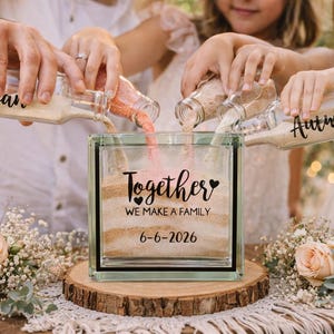 May include: A clear glass sand ceremony block with the words "Together We Make a Family" and the date 6-6-2026. Three bottles pour sand into the block. The bottles are labeled with names. The block sits on a wooden slice.