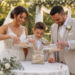 May include: A wedding sand ceremony with a bride, groom, and child pouring sand into a glass container. The container has the words "Together Forever" and names. The bride wears a white lace dress, and the groom wears a tan suit.