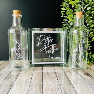 May include: Two clear glass bottles with cork stoppers and personalized names "Paul" and "Brittany" and a clear glass box with the text "Better Together 03-29-2025" on a wooden surface.