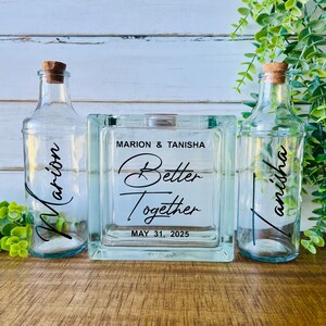May include: Two clear glass bottles with cork stoppers and a central square glass block. The left bottle features the name Marion, the right bottle features Tanisha, and the center block displays Marion & Tanisha Better Together MAY 31, 2025.
