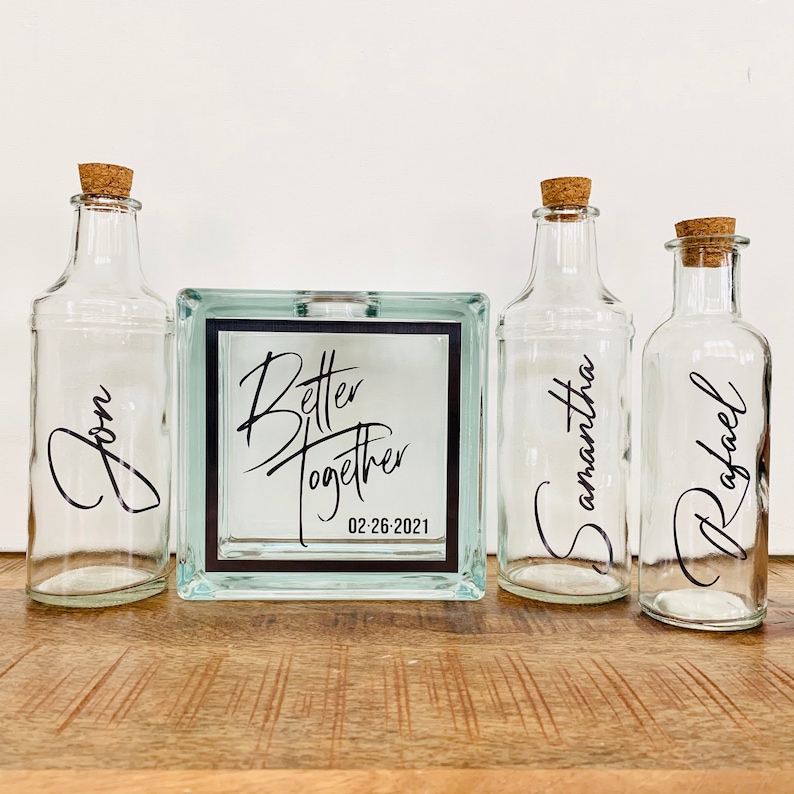 May include: Four clear glass bottles with cork stoppers and personalized labels. The bottles are arranged in a row on a wooden surface. The center bottle is in a square glass box with the text "Better Together 02-26-2021".
