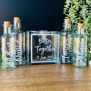 May include: Four clear glass bottles with cork stoppers and a square glass block. The bottles feature white script names: Aiden, David, Tamesia, and Ethen. The glass block displays white text reading Better Together and 12 June 2025.