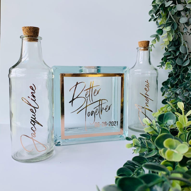 May include: Two clear glass bottles with cork stoppers and gold lettering. The bottles are labeled "Jacqueline" and "Andrew". A square glass box with a gold frame and the words "Better Together 06-26-2021" in gold lettering.