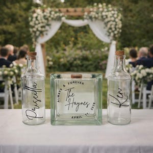 May include: A wedding sand ceremony set with two clear glass bottles and a glass block. The bottles are labeled "Priscilla" and "Ken". The block is inscribed with "Always & Forever The Haynes April 11, 2026". The set is on a white table.