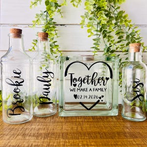 May include: Clear glass bottles and a glass block with cork stoppers. The bottles are labeled "Brookie", "Pauly", and "Sis" in black script. The glass block reads "Together WE MAKE A FAMILY 02.14.2026" in black.