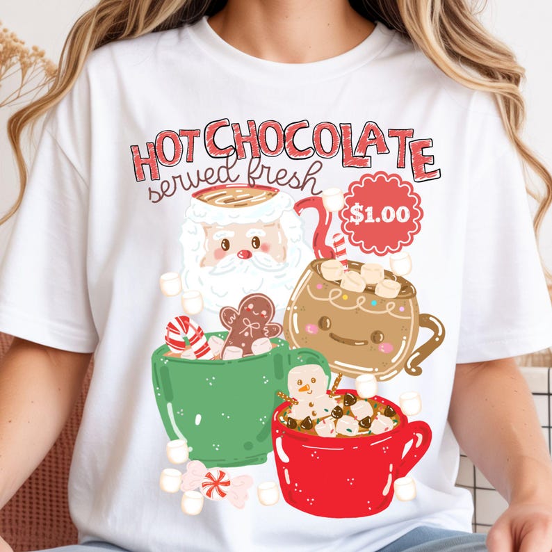 May include: A white t-shirt featuring a graphic print with the text "HOT CHOCOLATE served fresh" and "$1.00". The design includes a Santa Claus mug, a brown mug, a green mug, a red mug, marshmallows, a gingerbread man, and a candy cane.