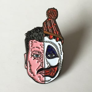 May include: A black and white enamel pin depicting a split face, half a man with a mustache and half a clown with a red smile. The clown is wearing a red and white hat with a red pom-pom.