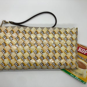 May include: A woven clutch bag made from recycled coffee packaging. The bag has a brown strap and a gold clasp. The bag is sitting on a white surface next to a packet of Kopiko Brown Coffee. The packet is yellow with brown text and a picture of a cup of coffee.