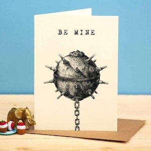 May include: A white card with the text "BE MINE" printed in black. A black and white illustration of a large, round, spiky object with a chain attached to it is in the center of the card.