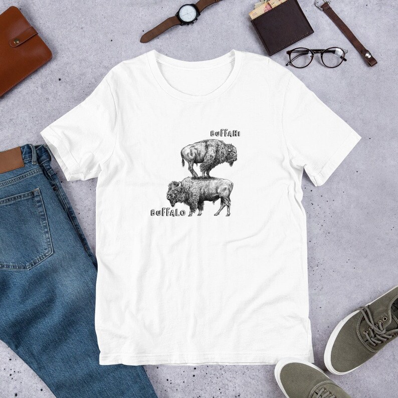 May include: White t-shirt with a black and white illustration of two buffalo stacked on top of each other. The top buffalo has the word "BUFFALO" written above it and the bottom buffalo has the word "BUFFALO" written below it.