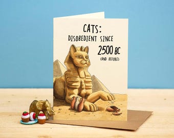 May include: Greeting card with a cartoon illustration of the Sphinx and pyramids. The card reads "CATS: DISOBEDIENT SINCE 2500 BC (AND BEFORE)". Miniature elephant and cupcakes are in front of the card. The background is a light blue wall and a wooden surface.