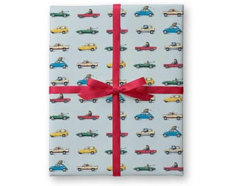 May include: A rectangular stack of light blue wrapping paper featuring a repeating pattern of various colorful vintage cars, including red, yellow, green, and blue models. The paper is secured with a bright red ribbon tied in a centered bow.