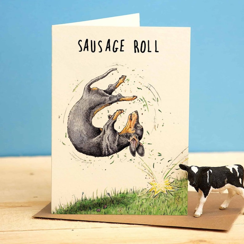 May include: A greeting card with a black and tan dachshund dog rolling in the grass. The card has the text "SAUSAGE ROLL" printed on it.