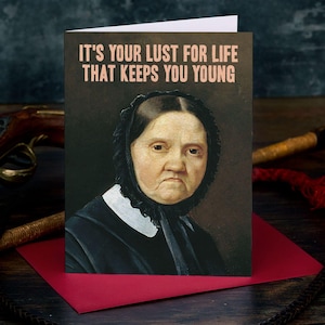 May include: A greeting card featuring a portrait of an elderly person wearing a black bonnet and a white collar. The text at the top reads, IT'S YOUR LUST FOR LIFE THAT KEEPS YOU YOUNG, printed in a light pink colour against a dark background.