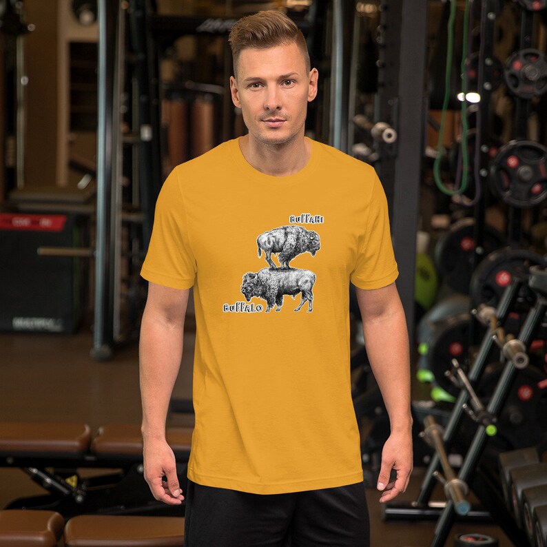 May include: A yellow short-sleeved t-shirt with a black and white illustration of two buffalo. The buffalo are stacked on top of each other. The top buffalo has the word "ruffian" written above it. The bottom buffalo has the word "buffalo" written below it.