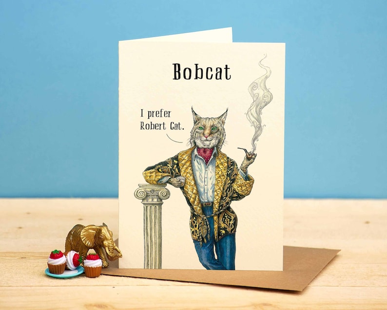 May include: Greeting card featuring a dapper bobcat with a pipe, wearing a patterned jacket and blue jeans. The card reads "Bobcat" at the top and "I prefer Robert Cat" below. A small gold elephant figurine and cupcakes are in the foreground.