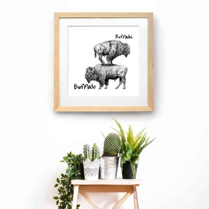 May include: A framed black and white print featuring two buffalo, one standing on top of the other. The word "Buffalo" is written below the bottom buffalo, and "Buffahi" is above the top buffalo. The frame is light brown.