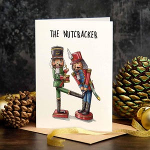 Op de afbeelding: Een witte wenskaart met de tekst THE NUTCRACKER en een illustratie van twee notenkrakerfiguren. De ene figuur draagt een groen uniform en bespeelt een trommel, terwijl de andere een rood-blauw uniform draagt en voor de eerste figuur buigt.