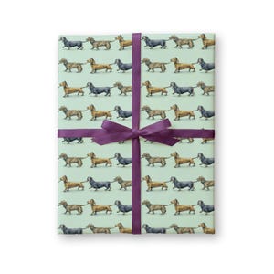 May include: A rectangular stack of wrapping paper featuring a repeating pattern of dachshund dogs in various colors including brown, black, and tan on a light mint green background, secured with a dark purple satin ribbon tied in a center bow.