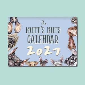 May include: A rectangular wall calendar with a light blue background featuring various dog illustrations around the border. The text reads The MUTT'S NUTS CALENDAR 2027 in dark grey and cream lettering, showcasing multiple dog breeds and ears.