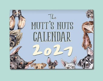 May include: A rectangular wall calendar with a light blue background featuring various dog illustrations around the border. The text reads The MUTT'S NUTS CALENDAR 2027 in dark grey and cream lettering, showcasing multiple dog breeds and ears.