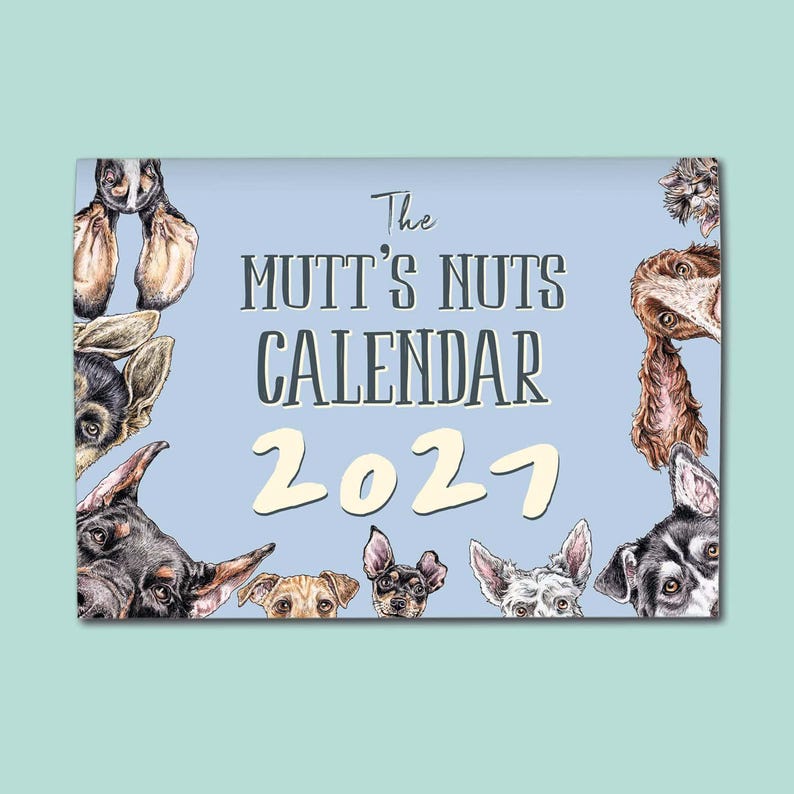May include: A rectangular wall calendar with a light blue background featuring various dog illustrations around the border. The text reads The MUTT'S NUTS CALENDAR 2027 in dark grey and cream lettering, showcasing multiple dog breeds and ears.