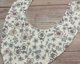 May include: A white bandana bib with a floral pattern in shades of pink and gray. The bib has a scalloped edge and a soft, absorbent texture.