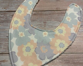 May include: A baby bib with a floral pattern in pastel colors. The bib features a U-shape design with a soft, absorbent fabric. The floral pattern includes light blue, peach, and yellow flowers on a white background.