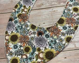 May include: A white cotton baby bib with a floral print featuring sunflowers, daisies, and other flowers in shades of yellow, orange, blue, and purple.