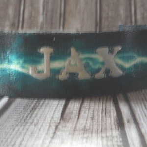 May include: A teal and black tie-dye fabric wristband with the name "JAX" embroidered in white.