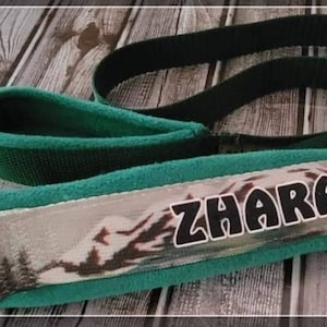 May include: A green and gray dog leash with the name "ZHARA" in bold black letters. The leash features a mountain landscape design and a padded handle for comfort. The leash has a silver metal ring.