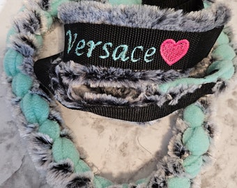 May include: A braided lead featuring a combination of mint green fabric and grey and black faux fur. A black nylon strap section is embroidered with the word Versace in light blue thread, accompanied by a small, bright pink embroidered heart.